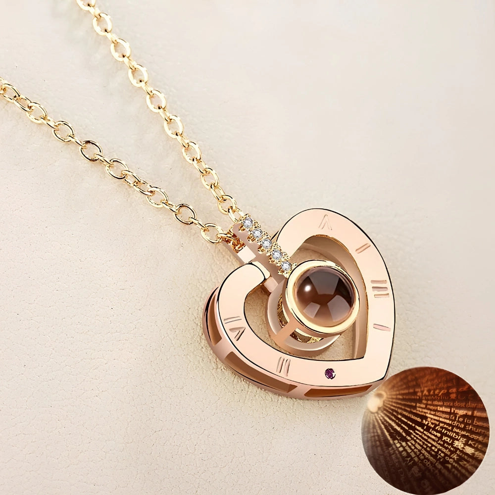 A gold heart-shaped pendant necklace with a Roman numeral design, a central brown gemstone, and a small inset circular detail showing a textured pattern, set against a light beige background.