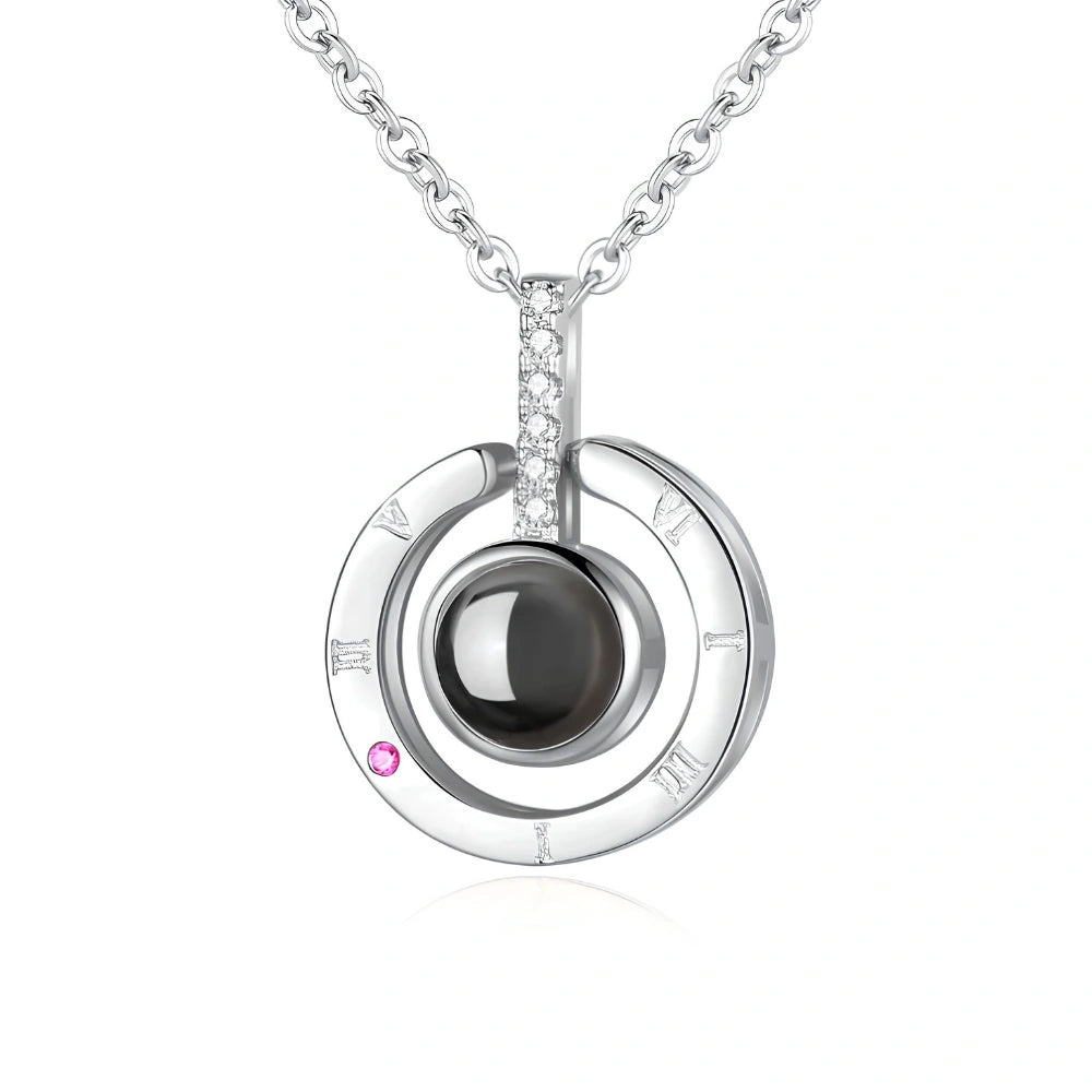 A silver pendant necklace featuring a double-ring design with a central dark gray sphere, a small pink gemstone, and a pavé-set clear crystal accent. The pendant is shown on a white background with a subtle reflection.