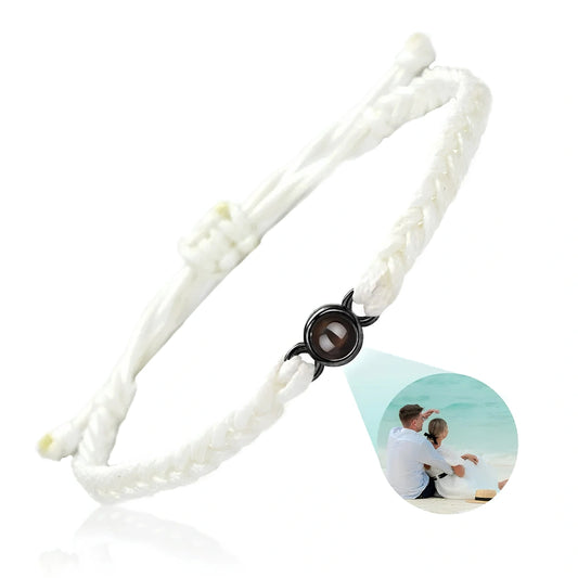 A white braided cord bracelet with a central black circular compartment containing a  photo projected of a couple by the sea, shown on a clean white background.