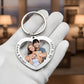 A silver heart-shaped keychain with a custom photo of a couple and engraved names and date, held in a white gloved hand against a blurred indoor background.