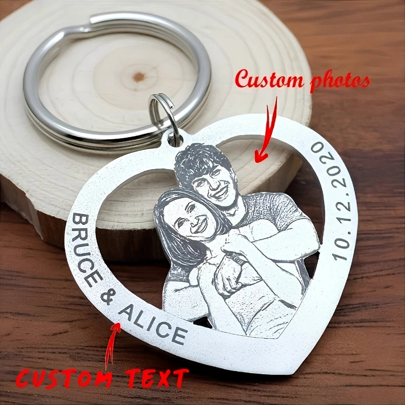 A heart-shaped silver metal keychain with engraved custom text and a custom photo of a couple, displayed on a wooden surface with a wood slice prop. 