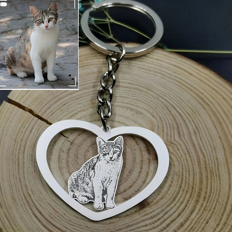 A silver heart-shaped keychain with an engraved cat design, shown on a wood slice background. A small inset photo in the top-left shows a real cat,  used to personalize the keychain.