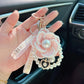 Hand holding a decorative keychain with a pink flower and pearl detail, set against a car interior background.